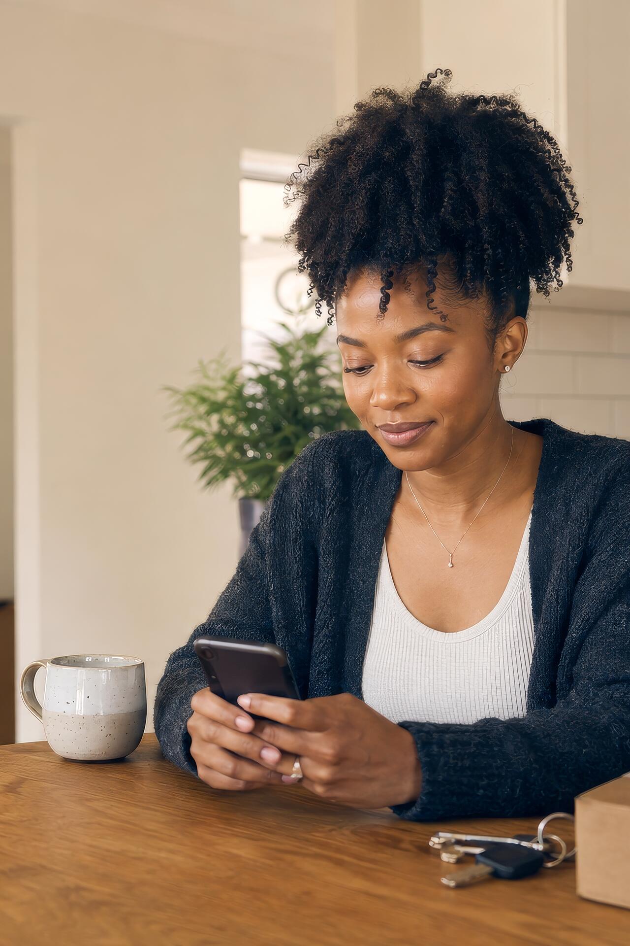A woman at a kitchen table with a mug, keys and a parcel, updating something on her phone